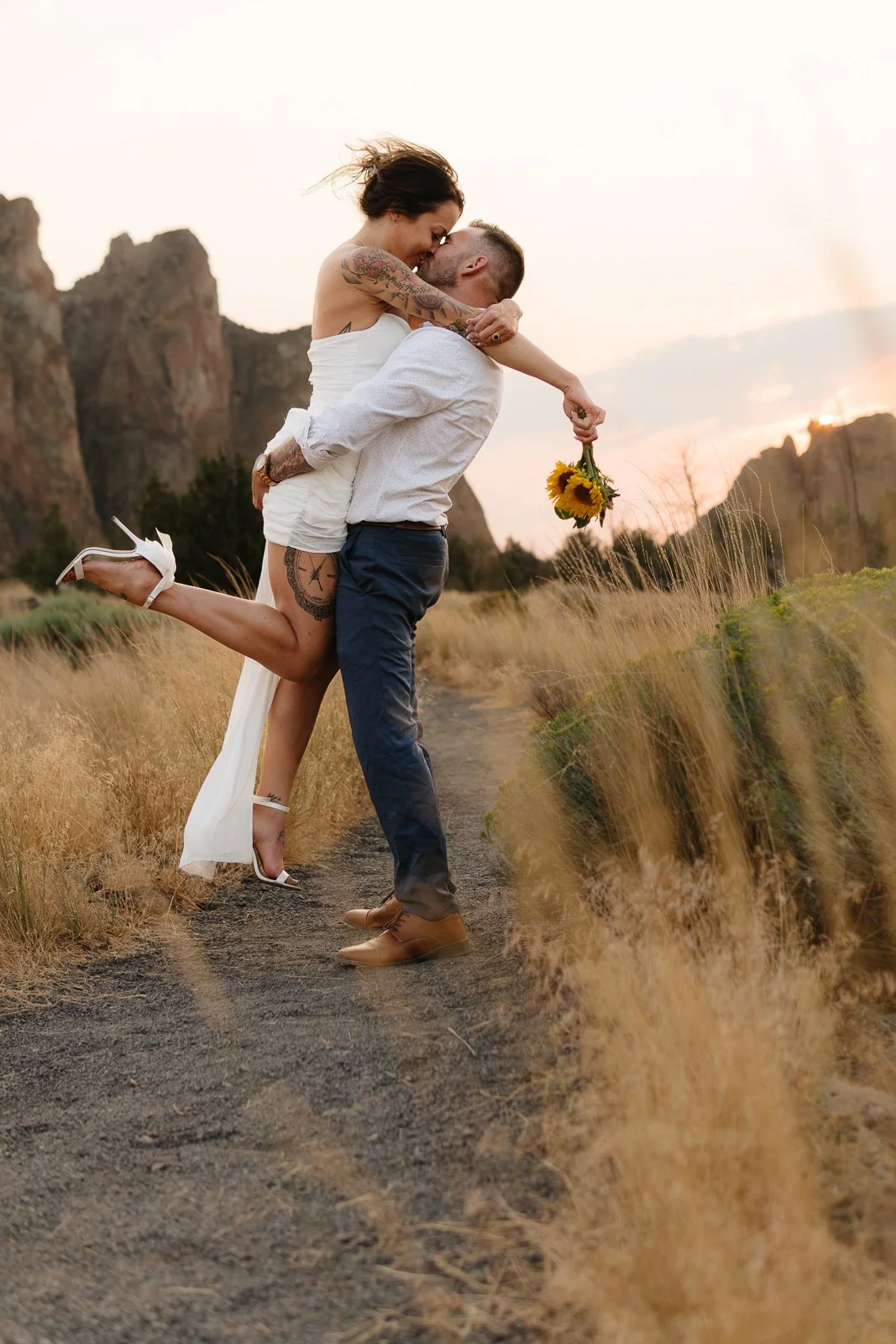  Jami and Matt share an intimate kiss at sunset in the heart of Smith Rock State Park, surrounded by golden grasses and iconic high desert cliffs. With Jami lifted off the ground, sunflower bouquet in hand and heels kicked up, the joy and passion between them radiates. The dramatic rock formations in the background glow with warm hues, capturing the raw beauty of Central Oregon and the wild, magnetic love they share. A perfect moment in one of Oregon’s most iconic landscapes. 